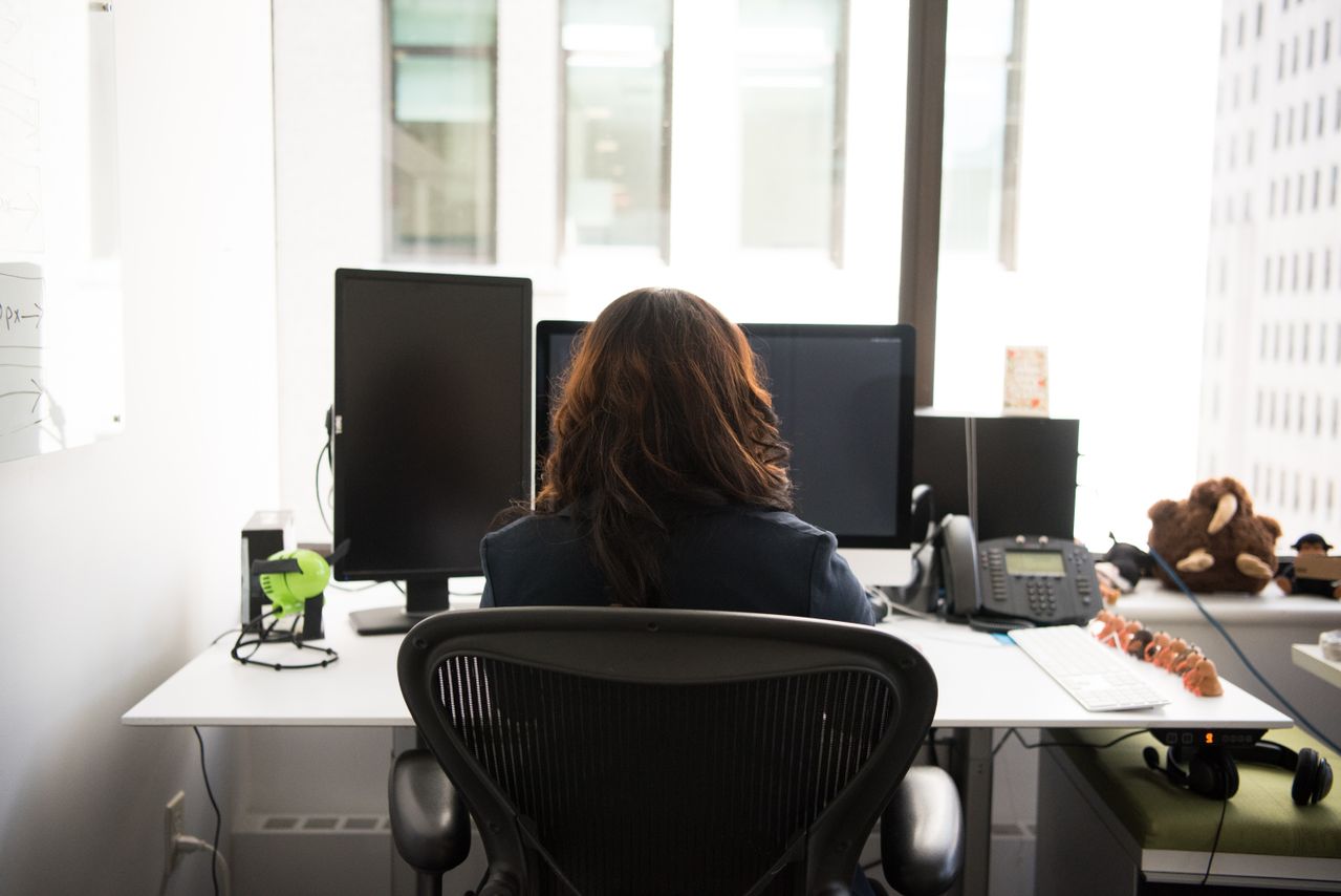 Donna vista di spalle, seduta su una sedia da ufficio davanti a due monitor